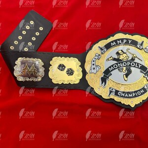 May include: A black and gold championship belt with a Monopoly theme. The belt features a silver Monopoly Man logo with the words "MONOPOLY CHAMPION" and "MNPY" on the buckle.