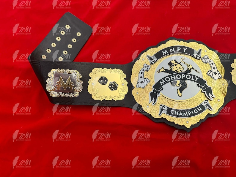 May include: A black and gold championship belt with a Monopoly theme. The belt features a silver Monopoly Man logo with the words "MONOPOLY CHAMPION" and "MNPY" on the buckle.