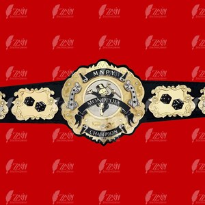 May include: A gold and black championship belt with a Monopoly board game theme. The center plate features a Monopoly Man figure and the words "MONOPOLY CHAMPION". The side plates feature dice and the letter "A" in a stylized font.
