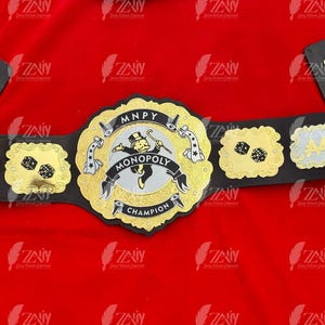 May include: A black and gold championship belt with a Monopoly theme. The center plate features a Monopoly man figure and the words "MONOPOLY CHAMPION".