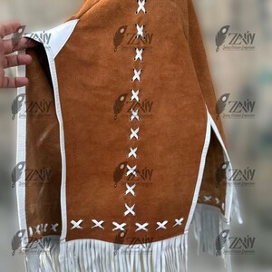 May include: A brown suede poncho with white trim and fringe. The poncho features white cross-stitch detailing and is trimmed with white leather. The bottom edge has a white fringe.