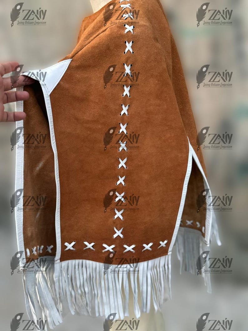 May include: A brown suede poncho with white trim and fringe. The poncho features white cross-stitch detailing and is trimmed with white leather. The bottom edge has a white fringe.