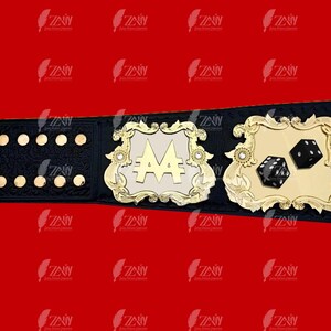 May include: A black and gold championship belt with a large, white, ornate plaque featuring the letter "A" with two dollar signs. The belt also has a smaller plaque with two black dice.