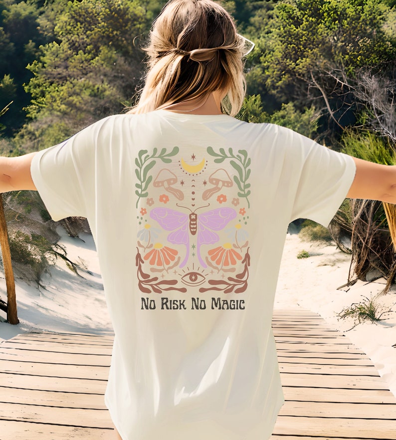 Op de afbeelding: Een wit T-shirt met een kleurrijk grafisch ontwerp met een vlinder, bloemen, paddenstoelen en een wassende maan. De tekst "No Risk No Magic" is onder het ontwerp gedrukt.