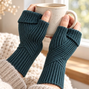 May include: A pair of dark teal fingerless gloves featuring a ribbed knit texture with a crisscross pattern across the back of the hand and a folded cuff at the top, held while grasping a speckled off-white ceramic mug.