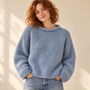 May include: A light blue long-sleeved sweater with a round neckline and a chunky knit texture. The garment features dropped shoulders and a relaxed fit, paired with blue denim jeans. The knit fabric has a soft, slightly fuzzy appearance.