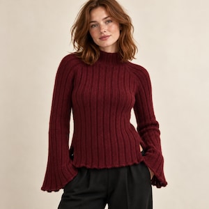 May include: A deep burgundy long-sleeve sweater featuring a ribbed knit texture, a mock neck collar, and flared sleeves. The garment has a straight hemline with side slits and is paired with plain black trousers.