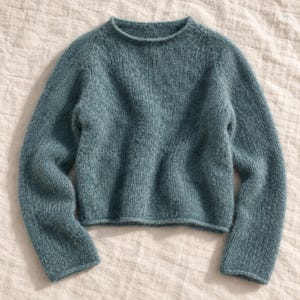 May include: A long-sleeved, crew-neck sweater in a muted teal blue color. The garment features a chunky, textured knit construction with a slightly cropped length and raglan sleeves, showcasing a consistent, fuzzy yarn appearance throughout.