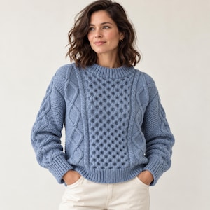 Könnte beinhalten: Ein hellblauer Langarmpullover mit einem markanten Zopfstrickmuster an den Ärmeln und einem strukturierten Wabenstrickmuster auf der Vorderseite, abgerundet durch einen gerippten Rundhalsausschnitt und gerippte Ärmelbündchen.