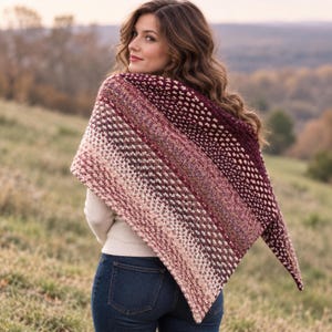 Pode incluir: Um xale de tricô triangular com um padrão de cor degradê que transita do bordô profundo e vermelho escuro para tons mais claros de rosa, creme e bege. O tecido texturizado exibe um padrão de ponto consistente em toda a peça.