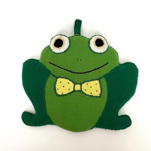 Może przedstawiać: Zielona pluszowa zabawka w kształcie żaby z żółtą muszką. Żaba ma duże, okrągłe oczy z czarnymi źrenicami i złotym obramowaniem. Zabawka jest wykonana z tkaniny i ma szyty wzór.