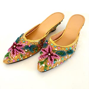 May include: A pair of gold-colored, pointed-toe mules with floral beadwork. The shoes feature a tan insole and a small heel. The floral design includes pink, green, and blue beads, with additional colorful embellishments.