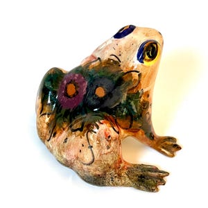Könnte beinhalten: Keramikfroschfigur mit einem einzigartigen, handbemalten Design. Der Frosch hat einen beigefarbenen Körper mit bunten floralen Akzenten in Lila, Grün und Orange. Seine Augen sind gelb mit blauen und schwarzen Details. Die Figur ist ein dekorativer Artikel.