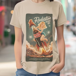 Puede incluir: Una camiseta beige con una ilustración de estilo vintage de una mujer montando una botella de Coca-Cola con forma de cohete. El texto "Radiation in every sip" está impreso en la ilustración.