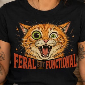 Può includere: Una t-shirt nera a maniche corte con la grafica di un gatto tigrato arancione con grandi occhi verdi e la bocca aperta che mostra i denti. Sotto il gatto, il testo recita FERAL BUT FUNCTIONAL in lettere maiuscole arancioni e rosse con stelle.