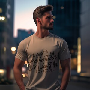 Puede incluir: Una camiseta beige claro con un dise&ntilde;o gr&aacute;fico de monta&ntilde;as y &aacute;rboles. El texto "Adventure Awaits" est&aacute; escrito en una fuente cursiva.