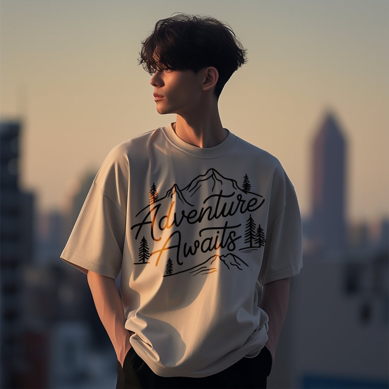 Puede incluir: Una camiseta beige con un estampado gr&aacute;fico negro de monta&ntilde;as y &aacute;rboles. El texto "Adventure Awaits" est&aacute; impreso en una fuente estilizada.