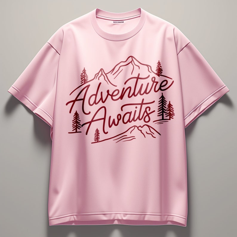 Puede incluir: Camiseta rosa de manga corta con un dise&ntilde;o gr&aacute;fico rojo de monta&ntilde;as y &aacute;rboles. El texto "Adventure Awaits" est&aacute; impreso en rojo en la camiseta.