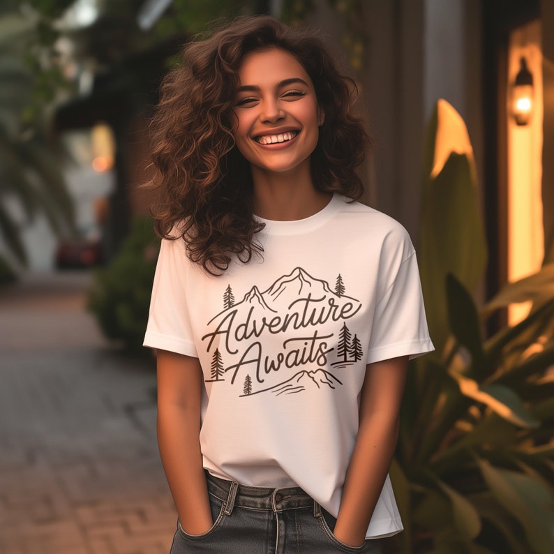 Puede incluir: Una camiseta blanca con un dise&ntilde;o gr&aacute;fico negro de monta&ntilde;as y &aacute;rboles. El texto "Adventure Awaits" est&aacute; escrito en una fuente cursiva.