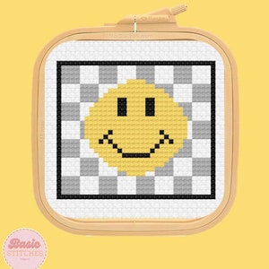 May include: A yellow smiley face cross-stitch pattern on a white background with black and gray checkered squares. The pattern is in a wooden embroidery hoop.