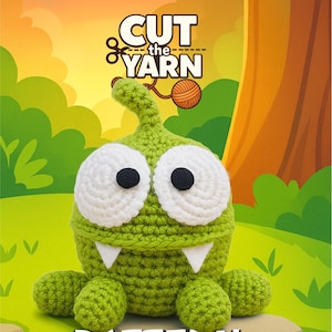 Puede incluir: Un monstruo amigurumi verde tejido a ganchillo con grandes ojos blancos y pupilas negras. El monstruo tiene pequeños dientes triangulares blancos y cuatro pequeños pies redondos. Se muestra el texto "CUT the YARN" y "PATTERN AMIGURUMI".