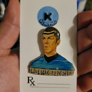 May include: A blue and white enamel pin featuring a portrait of a man with the text "LIVE LONG & PROSPER" on a banner below. The pin is on a white card with a blue circle and the text "Klonopins".