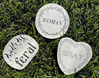 May include: Three white round stickers with black designs. The first sticker has a barbed wire circle with the word "WOMAN" inside. The second sticker has a flame design with the word "feral" inside. The third sticker has a heart shape with a chain border and the word "BRAT" inside.