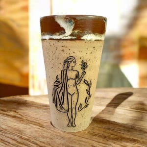 Puede incluir: Una taza de cerámica con un borde marrón y un cuerpo beige moteado. Una ilustración en blanco y negro de una mujer sosteniendo una rama con hojas está impresa en la taza.