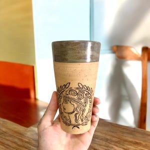May include: A ceramic mug with a brown and green glaze. The mug features a black and white design of a woman's face with a wreath of leaves around it.
