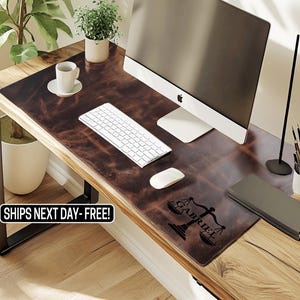 May include: A brown leather desk mat with a scale of justice graphic and the name "GABRIEL". The mat is on a wooden desk with a computer, keyboard, and mouse. A white coffee cup and plant are also visible.