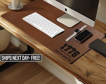May include: A brown leather desk mat with a white keyboard, computer, smart watch, and phone. The mat has the text "1776 - Land of the Free - Kevin Smith" printed on it. The desk is made of wood.