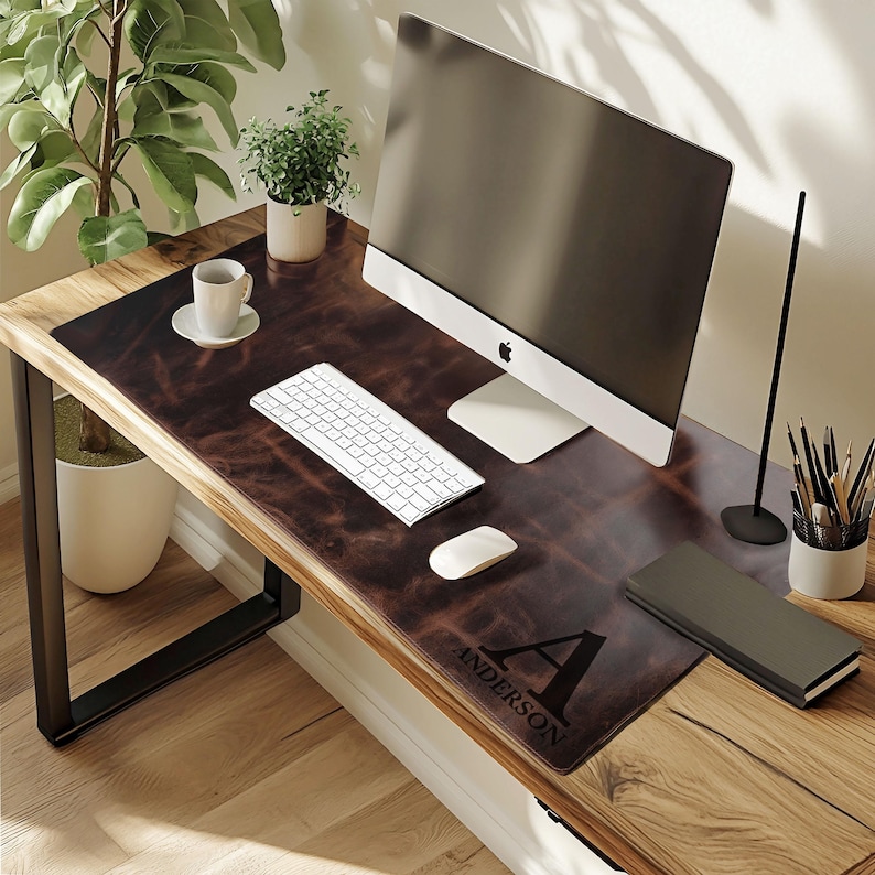 May include: A brown leather desk mat with the name "ANDERSON" and the letter "A" printed on it. A white keyboard, mouse, and computer monitor sit on the mat. A cup of coffee and a small potted plant are also on the desk.