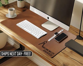 May include: A brown leather desk mat with a compass design and the text "William Miller Architect". A white keyboard, a white Apple Watch, and a black smartphone are on the mat.