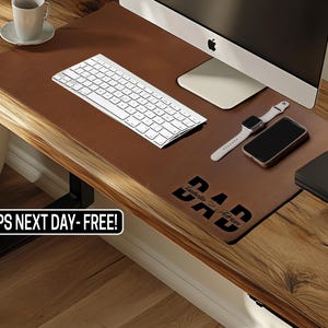 May include: A brown leather desk mat with a white keyboard, computer, smart watch, and phone. The mat has the word "DAD" in black, with a name below. The desk is made of wood.