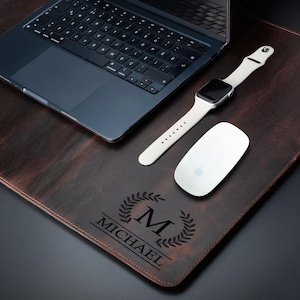 May include: A brown leather desk mat with a laptop, white mouse, and a smartwatch. The mat is personalized with the name "MICHAEL" and a letter "M" inside a laurel wreath design. The laptop is black.