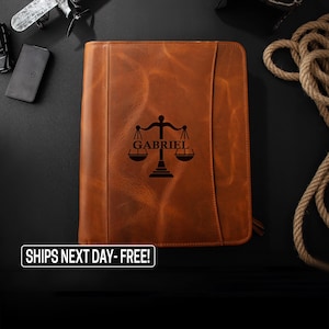 May include: A brown leather planner with a zipper closure. The planner has a scale of justice and the name "GABRIEL" engraved on the front.