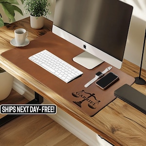 May include: A brown leather desk mat with a white keyboard, watch, and phone. The mat has a scale of justice graphic and the name "GABRIEL" etched on it. A computer monitor and a coffee cup are also visible.