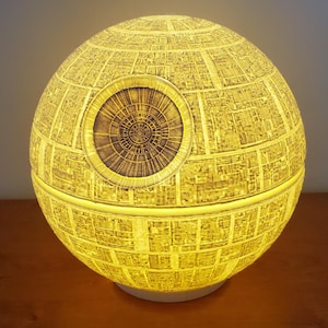 Peut inclure: Lampe sphérique jaune, évoquant l'Étoile de la Mort de Star Wars. La surface est détaillée avec une zone circulaire en retrait. Elle repose sur une base blanche et émet une lueur chaude, créant un objet décoratif sur le thème de la science-fiction.