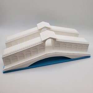 Może przedstawiać: Biały plastikowy model Mostu Rialto w Wenecji, Włochy. Most jest łukowy i ma szczeg&oacute;łowy projekt. Model stoi na niebieskiej podstawie.
