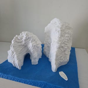 May include: A miniature diorama featuring two textured white iceberg formations on a blue base, with a small white boat. The scene evokes a polar landscape, suitable for model enthusiasts or decorative purposes.