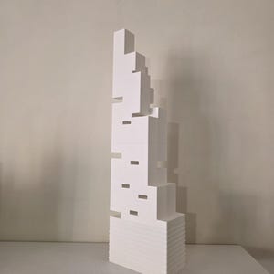 Peut inclure: Une sculpture abstraite blanche avec un design en couches. La sculpture a une base rectangulaire et une série de formes géométriques empilées. De petites découpes rectangulaires sont visibles sur les côtés. L'œuvre est exposée sur une surface blanche.