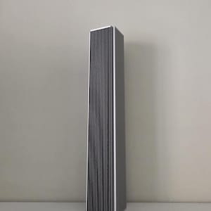 May include: A tall, rectangular speaker with a silver frame and a black, vertically-lined front panel. The speaker is standing upright on a white surface against a light grey wall. The design is minimalist and modern.