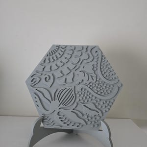 Könnte beinhalten: Eine graue, sechseckige dekorative Fliese mit einem floralen und geometrischen Reliefmuster. Die Fliese wird von einem passenden grauen Ständer mit gebogenen Beinen getragen. Das Design zeigt stilisierte Blätter, Ranken und gepunktete Texturen.
