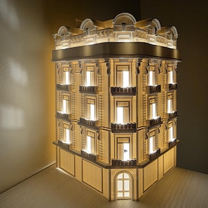 Può includere: Modello illuminato di un edificio con un design architettonico classico. Il modello presenta numerose finestre, finiture decorative e un bagliore caldo dall'illuminazione interna. L'edificio è una struttura angolare, che mostra la sua intricata facciata.