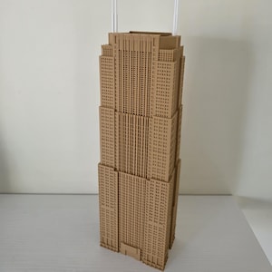 May include: A detailed model of a skyscraper, likely the Empire State Building, in a light brown color. The model has many windows and two white antennas at the top. The building is a multi-story structure with a rectangular base.