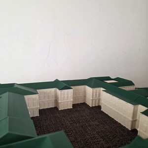 May include: An architectural model of a building, with white walls and green roofs. The model displays multiple sections, with a textured brown surface in the courtyards. The design includes rectangular windows and a symmetrical layout.