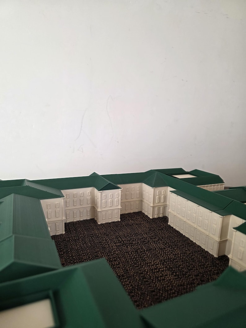May include: An architectural model of a building, with white walls and green roofs. The model displays multiple sections, with a textured brown surface in the courtyards. The design includes rectangular windows and a symmetrical layout.