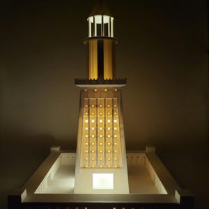 May include: A white model of a lighthouse with a light on at the top. The lighthouse is on a white base with a square shape.