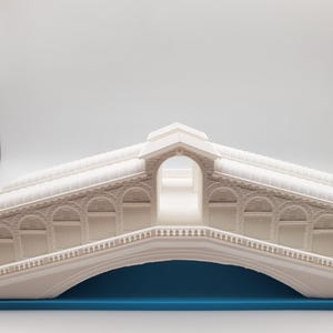 Może przedstawiać: Biały model mostu Rialto w Wenecji, Włochy. Most jest wykonany z plastiku i ma szczeg&oacute;łowy projekt. Most stoi na niebieskiej podstawie.