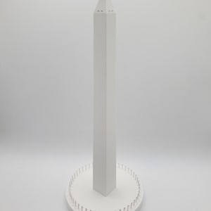 May include: A white, geometric sculpture resembling a tall obelisk. The structure has a pointed top and a rectangular base with small, evenly spaced pegs around its perimeter. The piece is set against a plain white background.